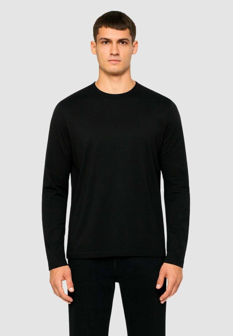 Maglietta nera a maniche lunghe realizzata in tessuto morbido, con scollatura rotonda e un branding discreto sul petto, indossata con pantaloni neri.