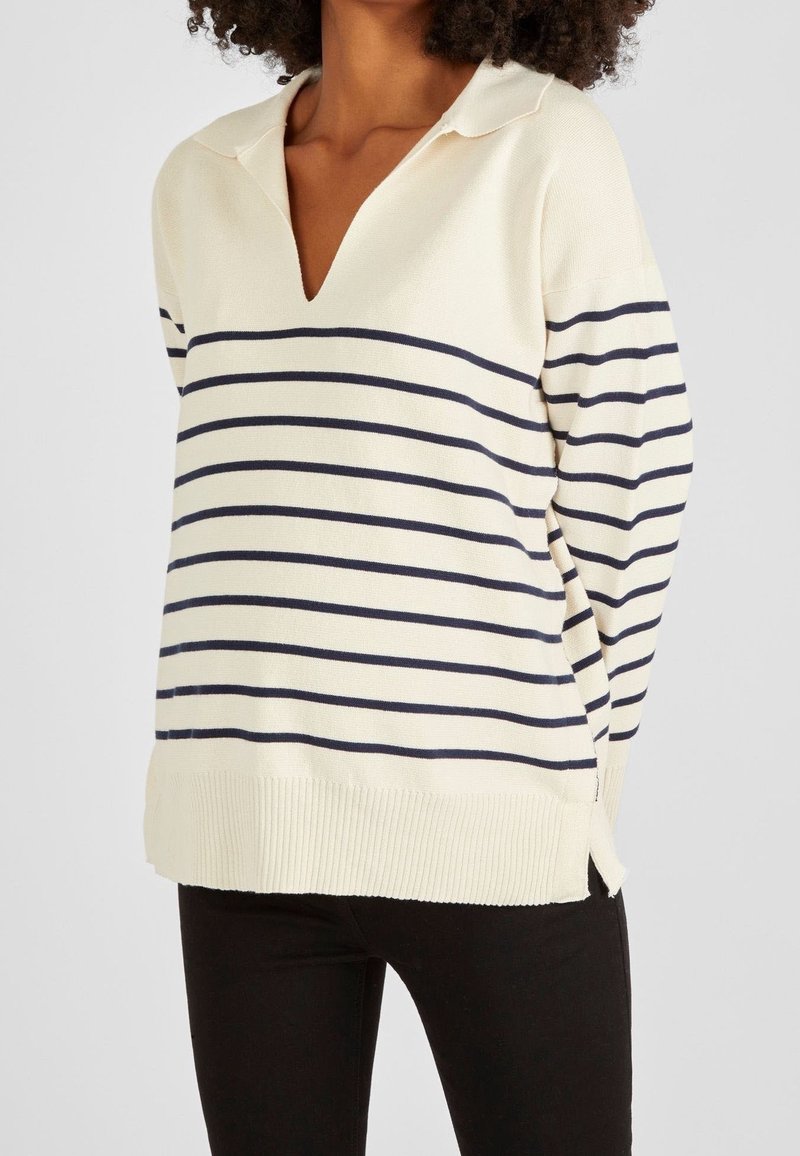 Femme portant un pull crème avec des rayures horizontales bleu marine, un col en V et un pantalon noir, debout devant un fond uni.