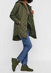 Olivově zelená prošívaná bunda s kapucí, postranními zipovými kapsami a dlouhým lemem, kombinovaná s modrými džíny a tmavě zelenými šněrovacími botami.