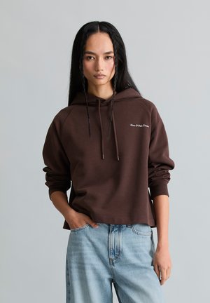 Kvinde med langt sort hår iført en brun hoodie og lyseblå jeans, stående med den ene hånd i lommen foran en ensfarvet baggrund.