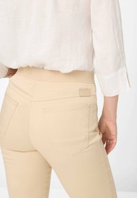 Beige high-waisted pants with a stretchy waistband and two rear pockets. Smooth texture, fitted design, and minimal stitching details.