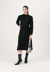 Black dress with long sleeves, ribbed texture, cinched waist with a belt. Paired with shiny black pointed-toe boots and a silver handbag.