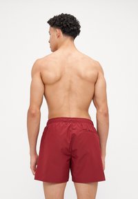 Shorts de bain bordeaux avec une taille élastique et une poche arrière. Fabriqués en tissu léger et soyeux. Le modèle est présenté de dos.
