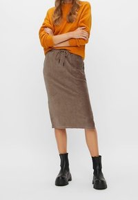 Brun kordflors midi kjol med dragsko i midjan, tillsammans med en orange stickad tröja och svarta ankelstövlar. Slät textur och avslappnad passform.