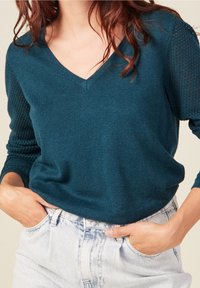 Femme portant un pull à col en V couleur sarcelle avec un détail en maille sur les manches et un jean délavé clair, les mains dans les poches avant.