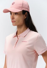 Vaaleanpunainen poolopaita, jossa on kaulus ja kolmen napin suljenta. Paita sisältää hienovaraisen logon samassa värissä. Yhdistetty yhteensopivan vaaleanpunaisen lippiksen kanssa.