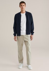 Man in een marineblauwe zip-up jas, wit t-shirt, beige broek en witte sneakers, staand tegen een effen lichte achtergrond.