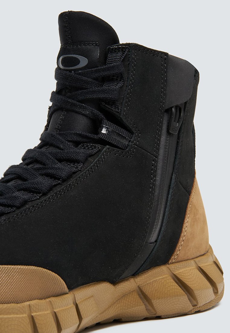 Bota de ante negra con una suela de goma texturizada en color marrón. Presenta un cierre lateral con cremallera y cordones planos negros. Diseño de caña alta para soporte del tobillo.