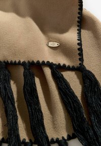 Beige fleece tørklæde med sorte kanter og frynser. Har et indsyet sølvlogo. Blød tekstur med dekorative kanter.