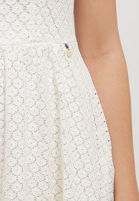 Robe en dentelle blanche avec un motif floral, jupette froncée et un petit accent décoratif avec des perles et un détail en pièce de monnaie métallique.