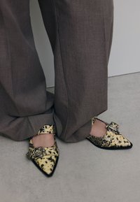 Schoen met spitse neus in een geel en zwart luipaardprint, voorzien van twee gespen en zilveren hardware, gecombineerd met grijze broek met wijde pijpen.