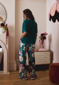 Zielona koszulka z krótkim rękawem w połączeniu z luźnymi spodniami w wzory, które mają kolorowe ilustracje deserów i kwiatów, umieszczone w stylowym wnętrzu.