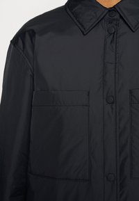 Veste noire à boutons avec col pointu et deux grandes poches frontales, fabriquée en tissu lisse et légèrement brillant.