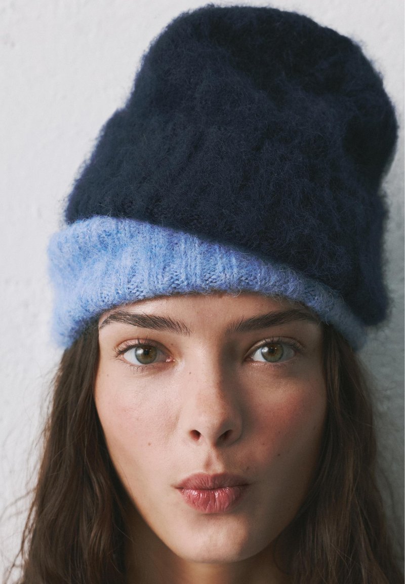 Jeune femme aux longs cheveux bruns portant un bonnet tricoté noir et bleu clair, regardant droit devant avec les lèvres légèrement pincées.