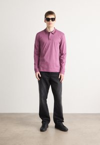 Camisa polo de manga longa roxa com colarinho, combinada com calças de denim pretas e sapatilhas pretas. O modelo usa óculos de sol escuros.