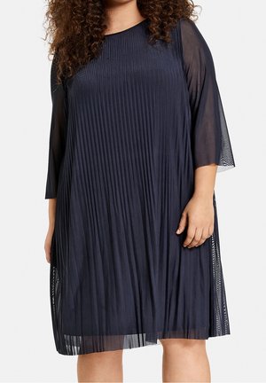 Kobieta w granatowej, plisowanej sukience do kolan z przezroczystymi rękawami o długości do łokcia, stojąca na jednolitym tle.