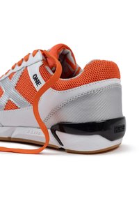 Zapatilla naranja y blanca con parte superior de malla, collar acolchado, suela de goma y un detalle en negro en el talón que contrasta. Cuenta con cordones planos de color naranja.