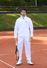 Baltas sportinis treningas su juodais akcentais, su užtrauktuku ir elastinėmis rankovėmis, derinamas su juodai baltais teniso bateliais. Laiko teniso raketę ir kamuoliuką.