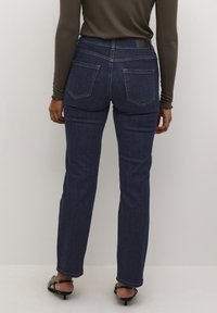 Jeans en denim bleu foncé avec une coupe droite, dotés de deux poches arrière et de coutures contrastées. Portés avec un haut à manches longues en côtelé.