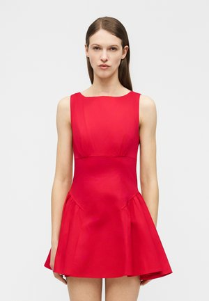 Mujer con cabello castaño lacio, lleva un vestido rojo ajustado sin mangas con falda acampanada, posando frente a un fondo claro y simple.