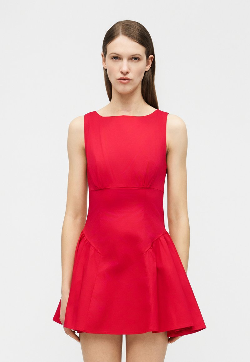 Γυναίκα με ίσια καστανά μαλλιά, φορώντας ένα αμάνικο εφαρμοστό κόκκινο φόρεμα με φουσκωτή φούστα, στέκεται μπροστά σε ένα απλό φωτεινό φόντο.