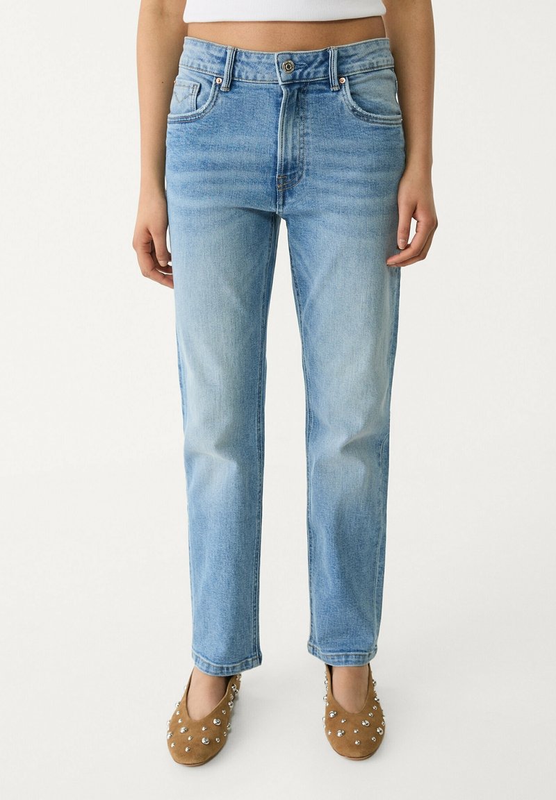 Femme portant un jean droit bleu clair associé à des chaussures plates marron cloutées, debout devant un fond uni.