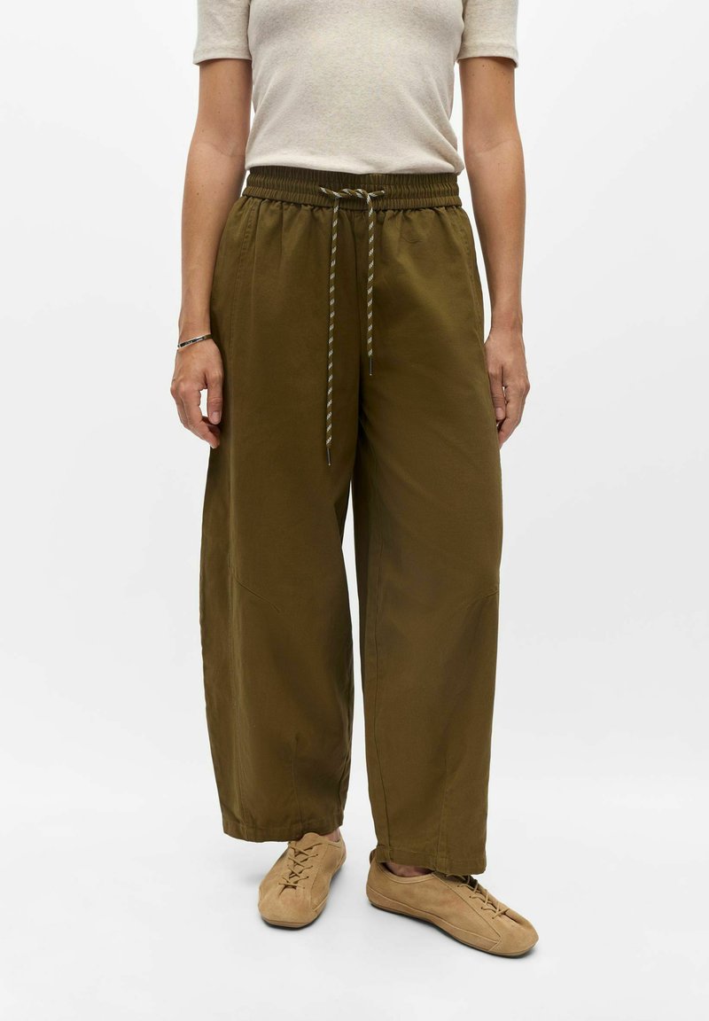 Personne portant un pantalon large vert olive avec taille à cordon, un haut beige à manches courtes et des chaussures décontractées marron clair à lacets sur fond blanc.