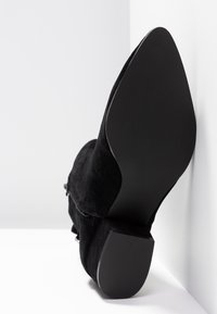 Bottine noire en daim avec bout pointu et talon bloc incliné vers le haut sur fond blanc en angle.