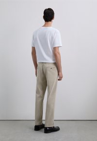Man facing away wearing a white short-sleeve shirt, beige pants with back pockets, and black dress shoes standing on a gray floor.