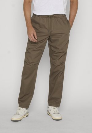 Mężczyzna w brązowych spodniach ze sznurkiem i białych tenisówkach, stojący na tle jasnego, jednolitego tła z rękami w kieszeniach.