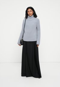 Sweater turtleneck a coste grigio chiaro, in vestibilità ampia, con maniche lunghe, abbinato a una gonna nera lunga fino al pavimento e a una piccola borsa a tracolla nera.