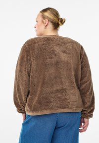 Veste en polaire marron avec une texture pelucheuse et des manches ballon, dotée d'un ourlet et de poignets côtelés, portée avec un pantalon bleu.