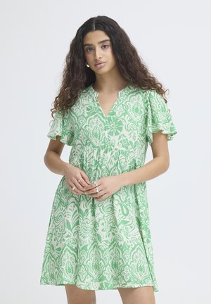 Junge Frau mit langem, lockigem Haar, die ein kurzes, grün-weiß gemustertes Kleid mit Blumenmuster trägt und die Hände gefaltet hält.