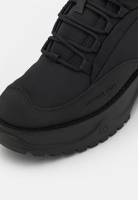 Botte noire imperméable mi-mollet avec une tige en tissu texturé, semelle en caoutchouc et un système de laçage proéminent arborant le logo de Michael Kors.