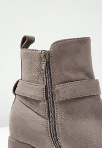 Primer plano de una bota de tobillo de ante topo con cremallera lateral y detalle de correa cosida sobre un fondo blanco.