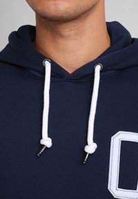 Sudadera azul marino con capucha, que cuenta con un cordón con extremos blancos, un gran parche blanco con contorno y una tela texturizada.