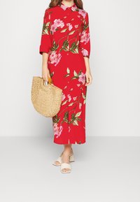 Abito floreale rosso con foglie verdi, chiuso con bottoni sul davanti e maniche a tre quarti. Abbinato a una borsa in paglia e sandali bianchi con tacco a blocco.