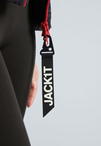 Schwarzes Stoffetikett mit weißer Aufschrift "JACK1T", befestigt an einem Reißverschluss mit roten Beschlägen. Der Hintergrund ist hellgrau und hat eine glatte Textur.