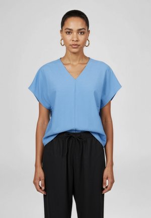 Frau mit goldenen Creolen, einer lockeren blauen V-Ausschnitt-Bluse mit kurzen Ärmeln und schwarzen Kordelzughosen, die vor einem schlichten grauen Hintergrund steht.