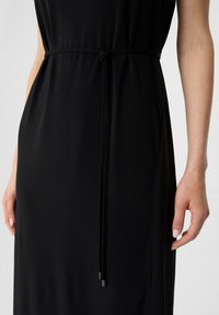 Schwarzes Kleid mit eingelötetem Taillengürtel, glatter Textur und subtiler Glanz. Verstellbarer Bindegürtel mit metallischen Enden.