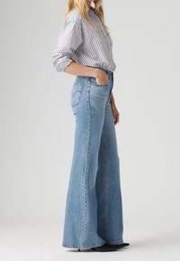 Jeans acampanados de cintura alta, de mezclilla azul clara con un acabado desgastado. Presenta dos bolsillos y un diseño distintivo en el bolsillo trasero.