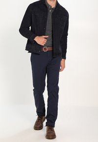 Mann in schwarzer Jacke, dunkelgrauem Poloshirt, marineblauen Hosen, braunem Gürtel und braunen Schnürschuhen, der vor einem schlichten Hintergrund nach vorne geht.