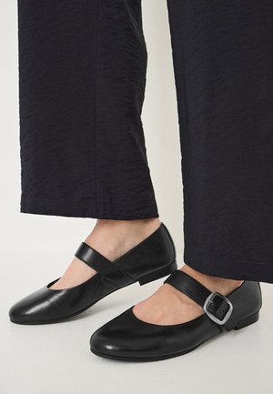 Voeten met zwarte leren Mary Jane schoenen met brede banden en een grote zilveren gesp, gecombineerd met gestructureerde zwarte cropped broek.