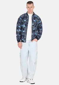 Marine bomberjack met een blauw flamingo- en palmbladenpatroon, geribbelde manchetten en zoom. Gedragen over een wit t-shirt, gecombineerd met lichte cargobroeken.