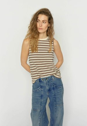 Naine, kellel on pikk laineline juuksed, kannab varrukateta beeži ja pruuni triibulist topi ning lahtisi siniseid teksapükse, seisab ühevärvilise valge tausta ees.