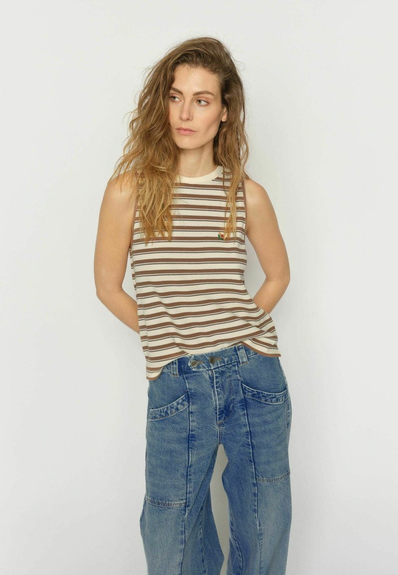 Hosszú hullámos hajú nő, aki ujjatlan bézs-barna csíkos felsőt és bő kék farmert visel, egyszerű fehér háttér előtt áll.