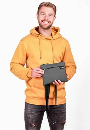 Šedé psaníčko se zipem, minimalistickým designem, připevněným popruhem a hladkou texturou, držené osobou v žluté mikině s kapucí.