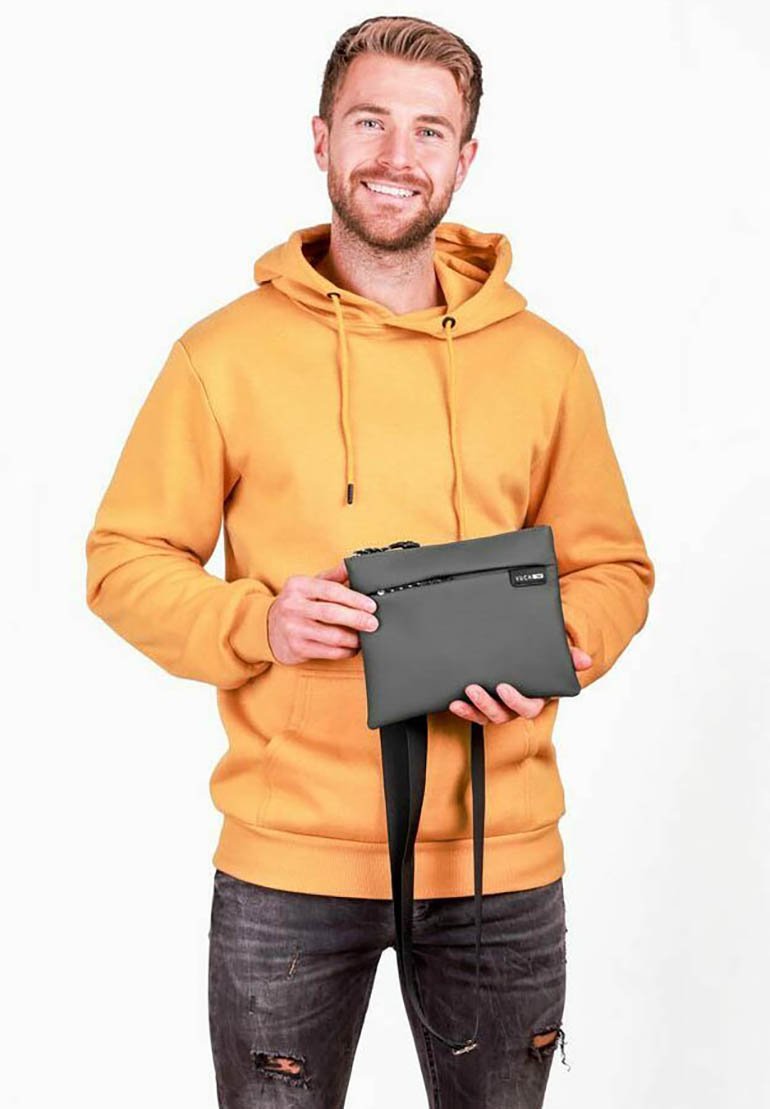 Šedé psaníčko se zipem, minimalistickým designem, připevněným popruhem a hladkou texturou, držené osobou v žluté mikině s kapucí.