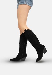 Schwarze kniehohe Stiefel aus Wildleder mit spitzen Zehen und dekorativen Nähten. Sie verfügen über einen Reißverschluss und einen mittelhohen Blockabsatz.