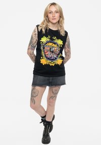 Black sleeveless cotton shirt with colorful floral graphic; paired with a denim skirt. Black chunky boots, sporting multiple tattoos.